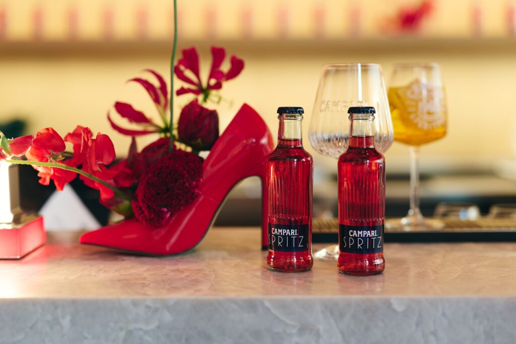 Zwei Flaschen Campari Spritz neben rotem High Heel und Blumendekoration